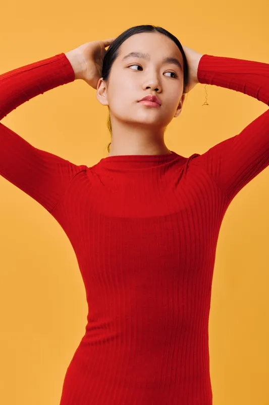 Young woman poses with hands behind head wearing red knit sweater against yellow background. Female model with natural makeup and calm expression in studio portrait.