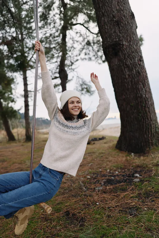 Outdoor scene with a smiling woman on a swing among trees in a park, wearing a cozy sweater and beanie, capturing carefree moments and a natural backdrop for lifestyle and seasonal leisure.