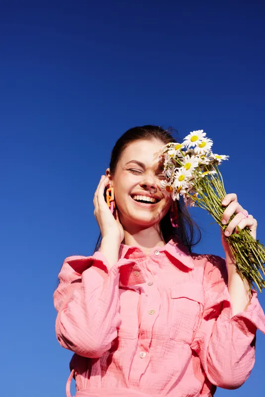 Happy young woman in a pink dress holding wildflowers against a vibrant blue sky, expressing joy and appreciation for nature and beauty