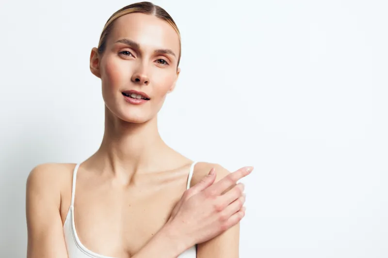 Portrait of young woman with clear skin wearing white tank top looking at camera with soft smile holding shoulder isolated on white background minimal beauty concept.
