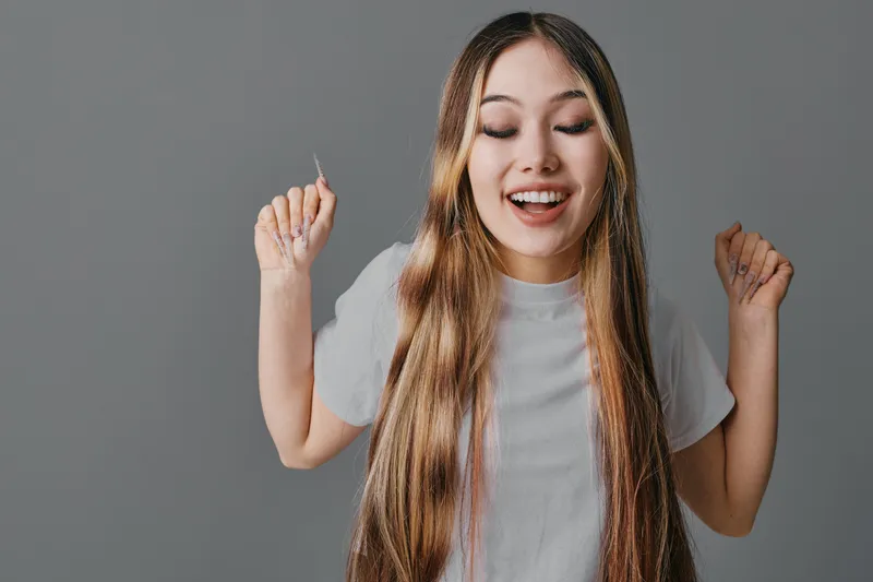 Young woman with long hair smiling and holding a small object in one hand on gray background. Casual female in white t shirt with emotional expression and closed eyes.
