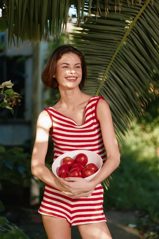 A cheerful woman in a red and white striped swimsuit holds a bowl of apples in a sunny outdoor setting. She smiles warmly, radiating health, joy, and vitality.