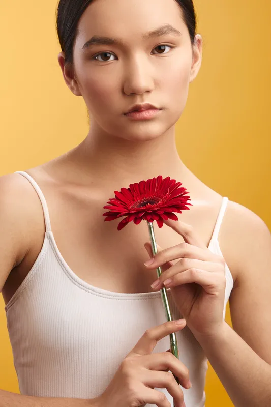 asian woman holding a red flower against a warm yellow backdrop, soft studio lighting highlights delicate features and a calm, poised expression in a contemporary portrait.