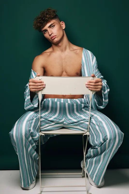 Young man with curly hair wearing striped pants and open shirt sitting on chair backwards against dark background. Fashion, casual style and confident pose in studio portrait.