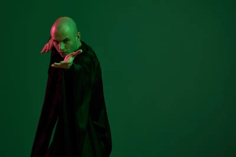Confident man posing dramatically in a dark garment against a green background, showcasing expressive body language. The image captures a strong sense of emotion.