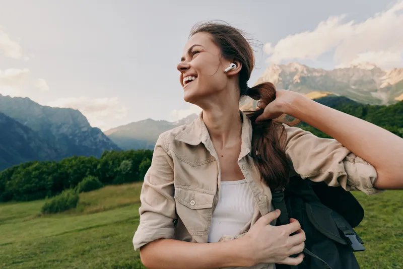 A young woman outdoors smiles while adjusting her backpack against a dramatic mountain backdrop, capturing carefree exploration.