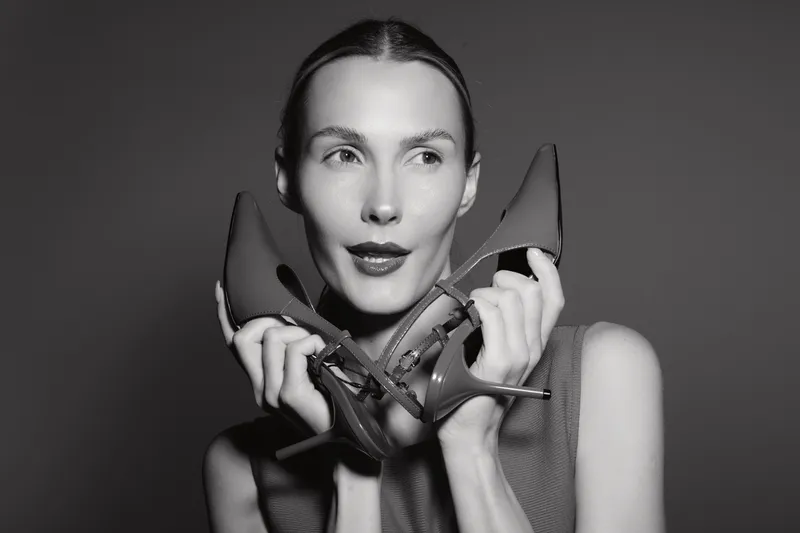 A young white woman with pale skin holds high heel shoes near her face, posing thoughtfully in monochrome studio lighting. Concept for fashion and style.