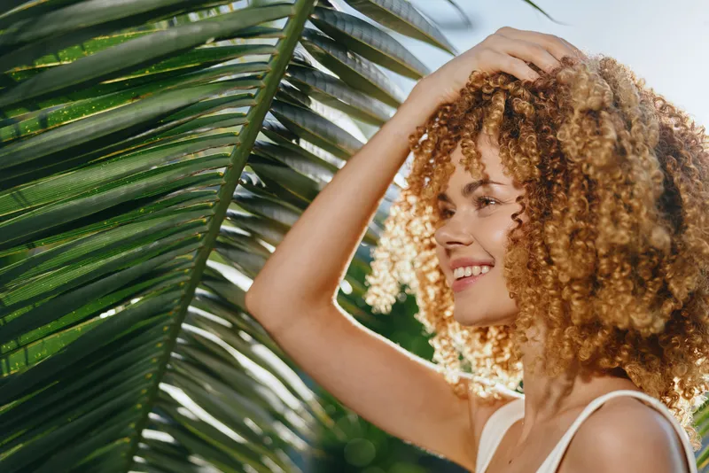 Smiling woman with curly hair posing outdoors near vibrant green palm leaves in bright natural sunlight, showcasing summer vibes and carefree happiness.