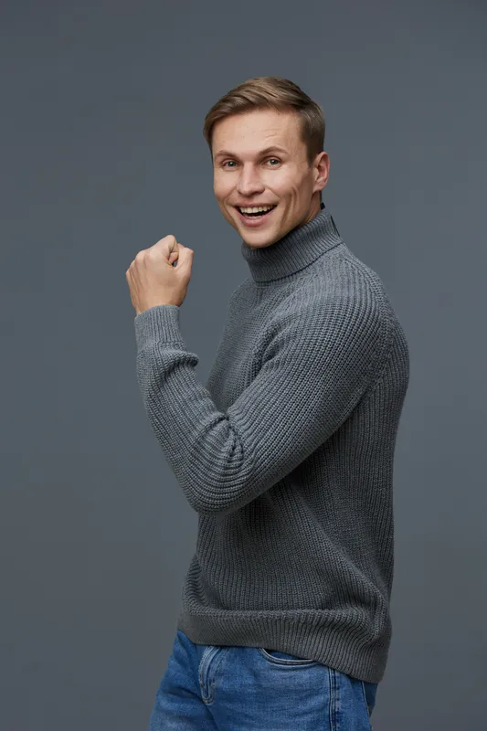 Happy cheerful young man in gray sweater looking at camera with joyful expression, fist raised, isolated on plain dark gray background, studio portrait, lifestyle concept.