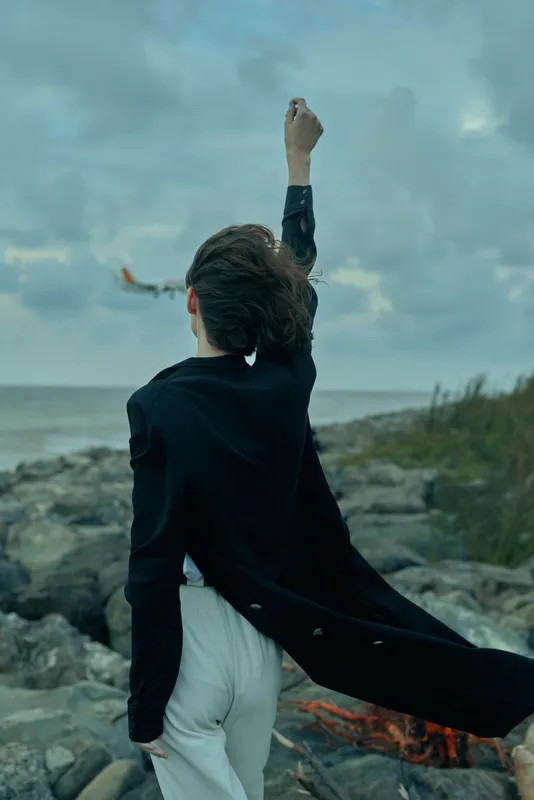 Victory stance on a rocky coast as a figure raises a fist, cape fluttering, ocean wind and dramatic silhouette convey triumph, empowerment, and bold determination.