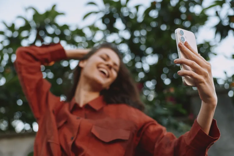A young woman in a red shirt takes a carefree selfie outdoors, hair tossed by the wind, capturing a candid moment of joy and confidence in a bright, natural setting