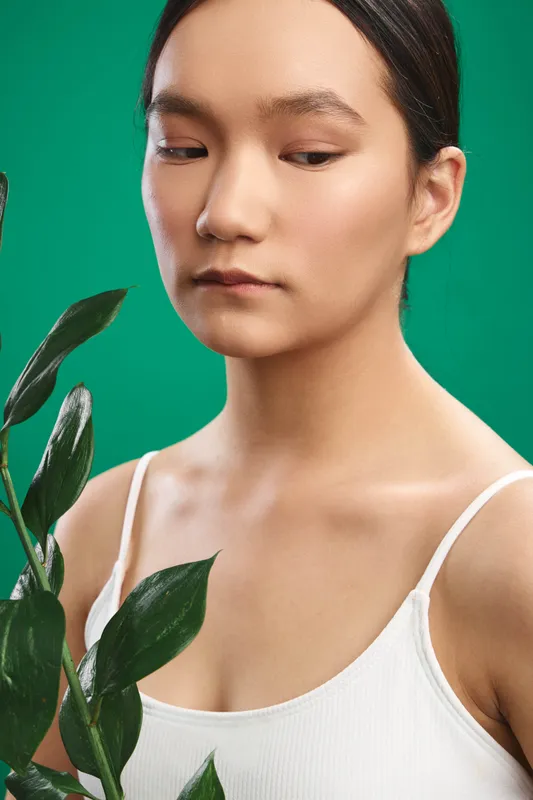 asian woman stands beside a leafy plant against a vivid green backdrop, wearing a white tank top, calm expression, soft lighting, minimal setting conveying natural simplicity.