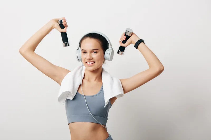 Young fit woman exercising with small dumbbells wearing sports bra and headphones, towel draped over shoulders, smiling against plain white studio background.