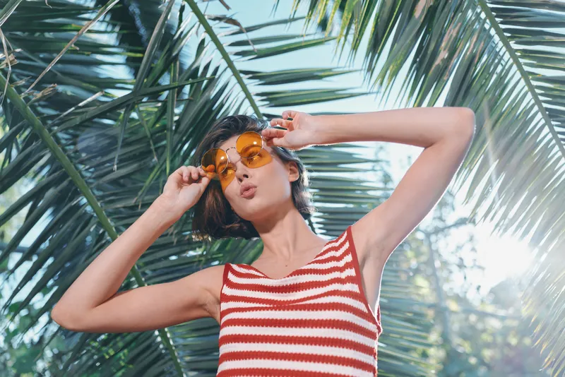 A stylish solo traveler poses under tropical palm leaves, wearing a red and white striped tank top and sunglasses. Bright sunlight creates a carefree summer vibe.