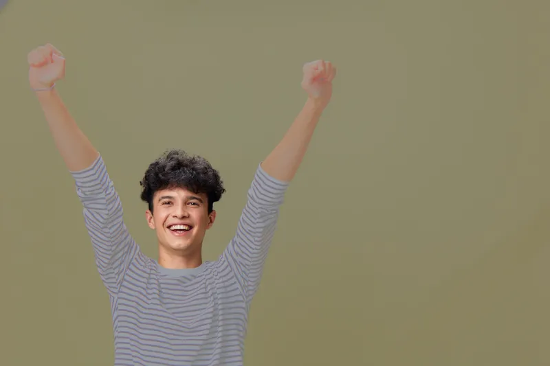 Happy young man celebrating success with raised arms wearing a striped shirt, expressing joy and confidence against a plain neutral background.