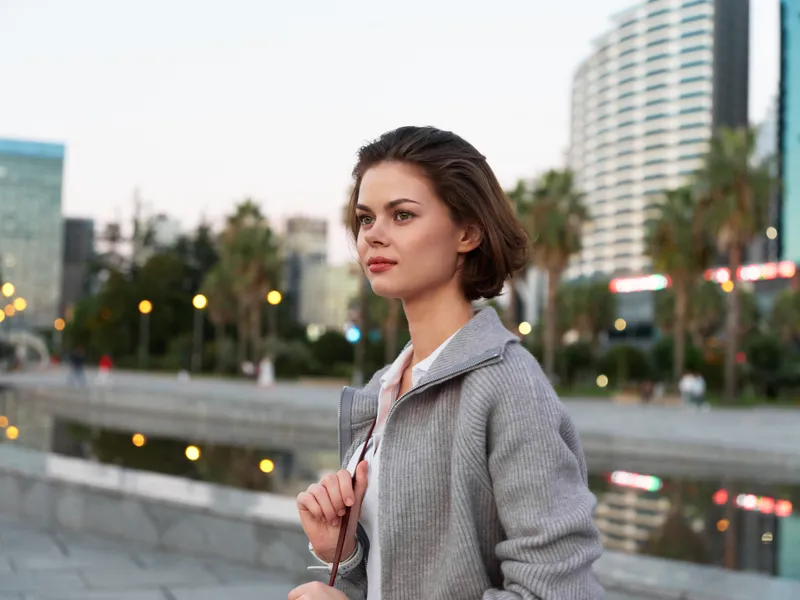 A stylish woman stands outdoors in an urban waterfront area, wearing a gray coat. She gazes ahead with confidence, exuding calm focus and a modern, professional vibe.