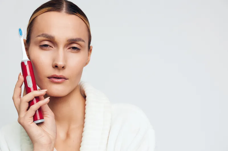 Young woman holds electric toothbrush near face with closed eyes in white robe isolated on light background showing dental hygiene concept and personal care routine at home.