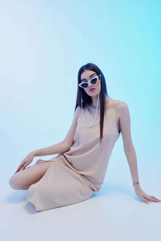 Young woman in a beige dress and white sunglasses poses seated on a light blue background. Concept of beauty, fashion, and modern style with elegant minimalism.