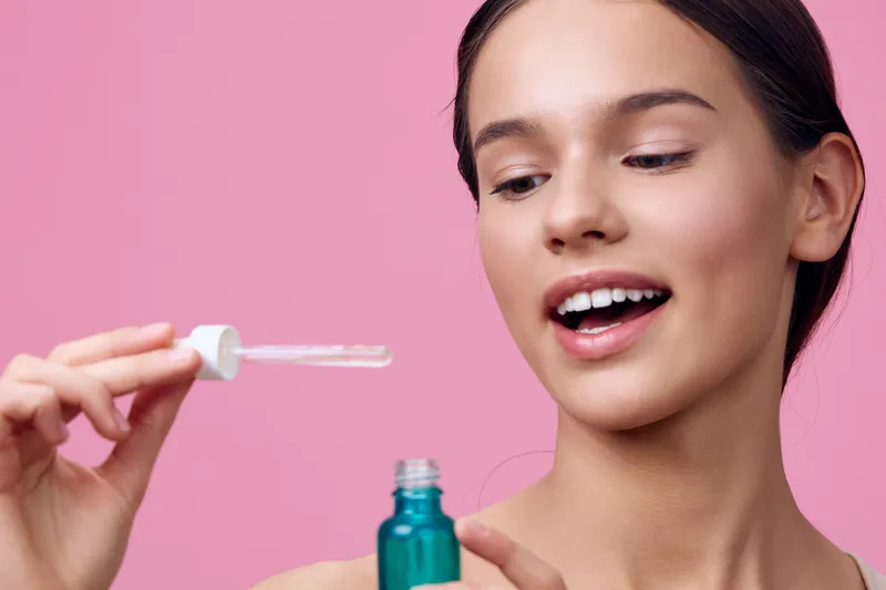 Young woman applying facial serum with a dropper, smiling, on a pink background emphasizing skincare routine and healthy lifestyle