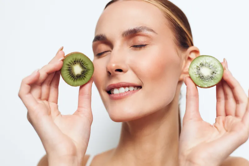 Young woman with closed eyes holding two halves of fresh kiwi fruit near her face. Concept of healthy eating, natural beauty, and fresh organic food. Bright studio lighting on white background.