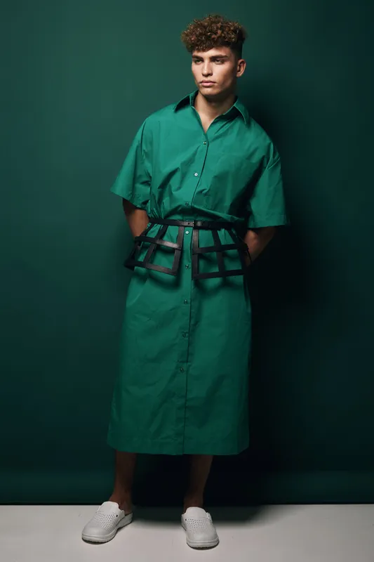 Young man standing in green oversized shirt dress with black harness belt and white sneakers against dark background. Fashion model posing with hands behind back in studio.