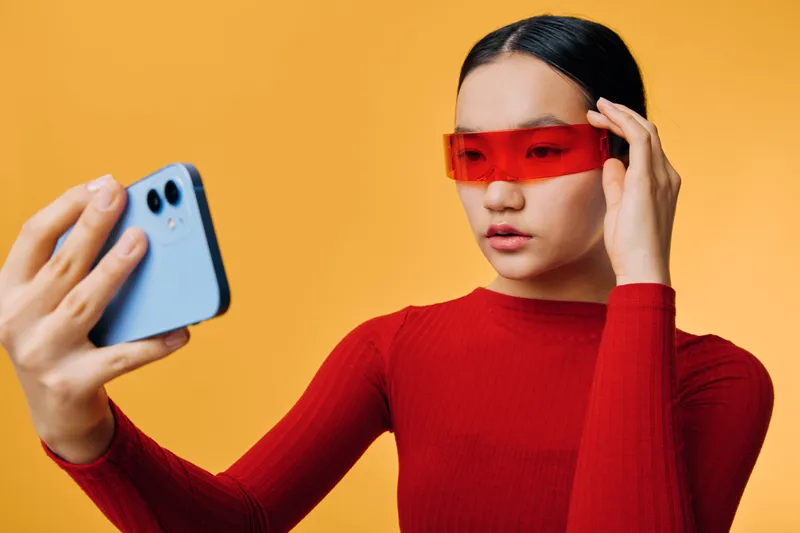 Young woman in red sweater and futuristic red glasses taking selfie with smartphone against yellow background. Fashion, technology, portrait and social media concept with minimal style.