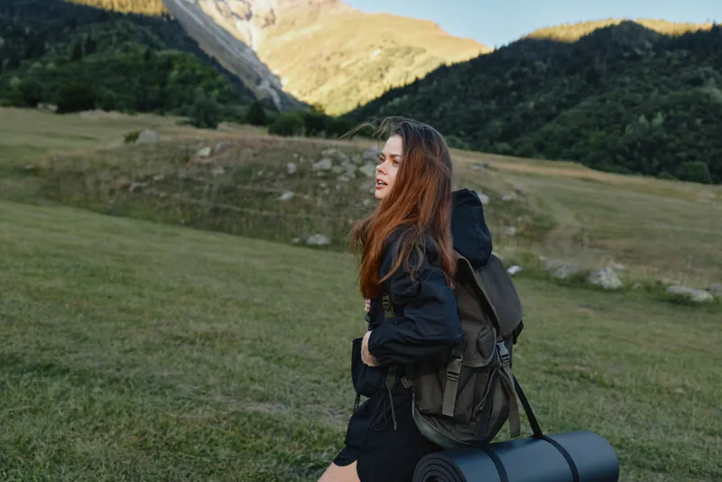 A cheerful traveler stands in a grassy meadow with a backpack, smiling as she gazes toward distant mountains, embodying energy, adventure, and an open outdoor spirit.