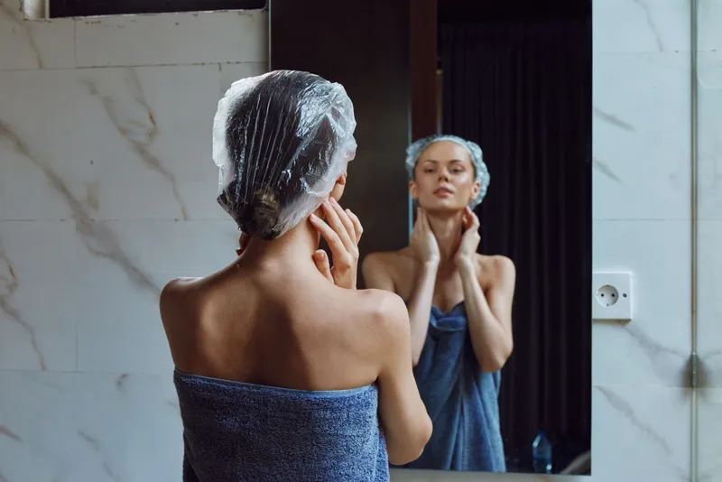 woman in shower cap and towel looking at mirror reflection in modern bathroom with white marble tiles, skincare routine, calm atmosphere
