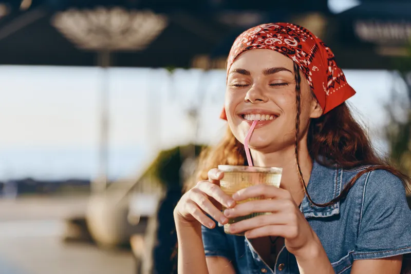 lifestyle woman enjoying drink in modern boho-western denim street style with film color tones, smiling outdoors under bright sunlight, relaxed and happy mood