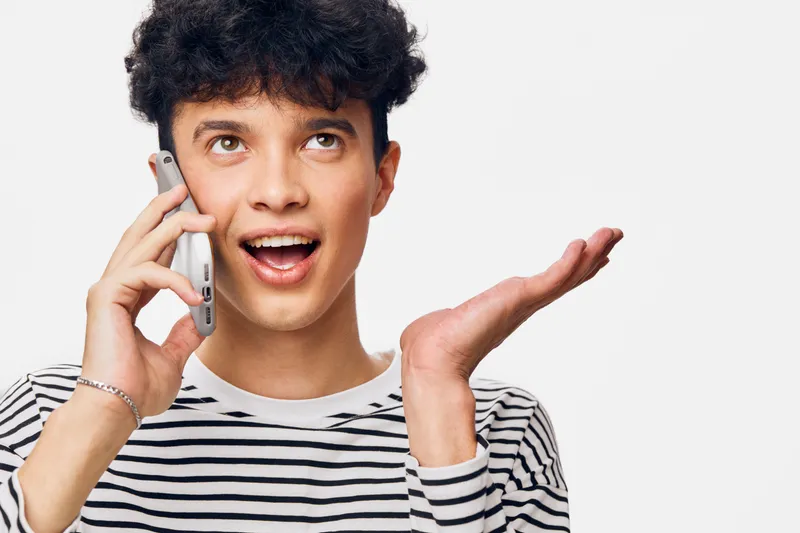 young man talking on smartphone with excited expression wearing striped shirt against white background, communication and technology concept