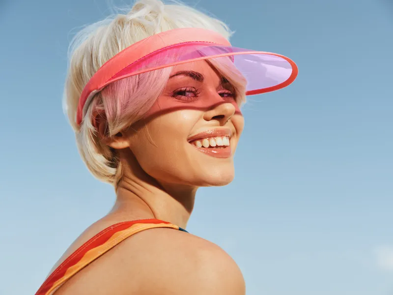 Close portrait of a smiling woman wearing a pink translucent visor outdoors under a bright sky, with vibrant colors and a confident, carefree mood.