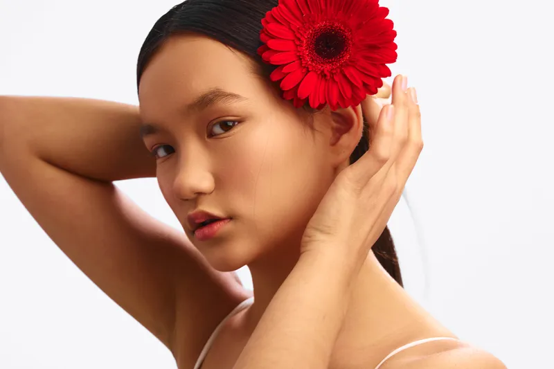 Asian woman with a red flower in her hair gazes softly at the camera in a studio portrait, presenting elegance, calm expression, soft lighting, clean background.