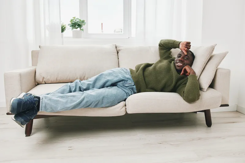 Young Black man relaxing on a cozy couch at home, wearing a green sweater and jeans, exuding a cheerful and relaxed mood in a bright interior