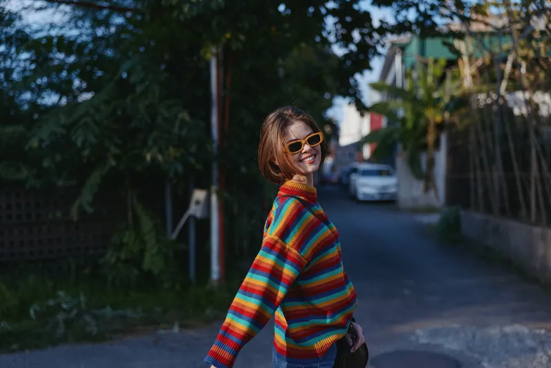 Woman in her early 20s, striped sweater and sunglasses smiling while turning on a sunlit suburban street; short hair, casual outfit, playful travel vibe and relaxed lifestyle.