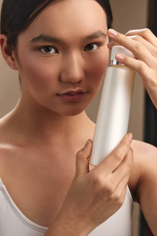 Asian woman holds a sleek cosmetic bottle near her face, highlighting a skincare routine in a calm studio with soft lighting and a focused, intimate mood.
