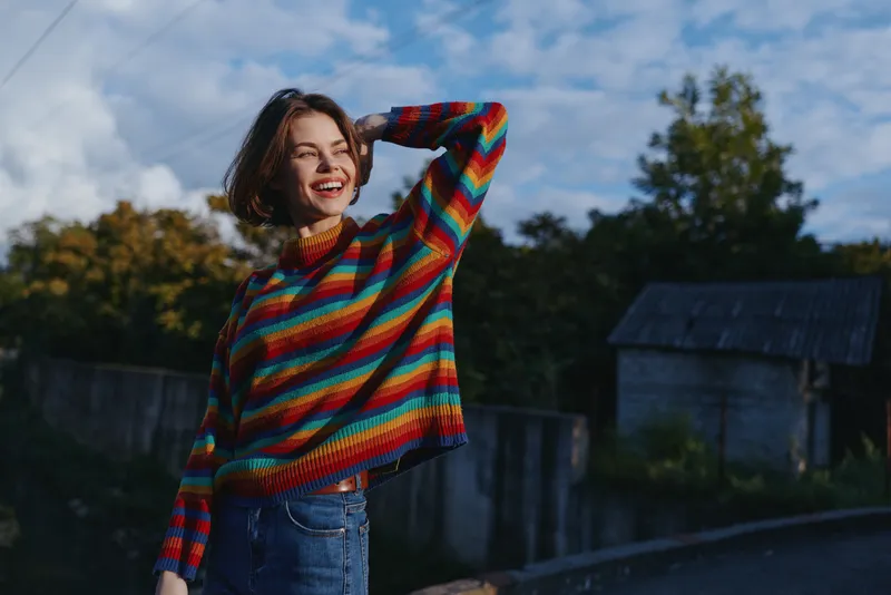 Young woman in a colorful striped sweater smiling woman outdoors in a rural village, short hair, casual outfit and jeans with hand on head, joyful portrait for lifestyle travel fashion.