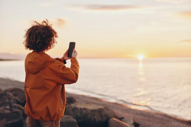 Woman lifestyle by sea at sunset, outdoors on rocky beach coast, taking a selfie with phone in hands, warm golden light, relaxed travel mood, rear view, calm waves and breeze.