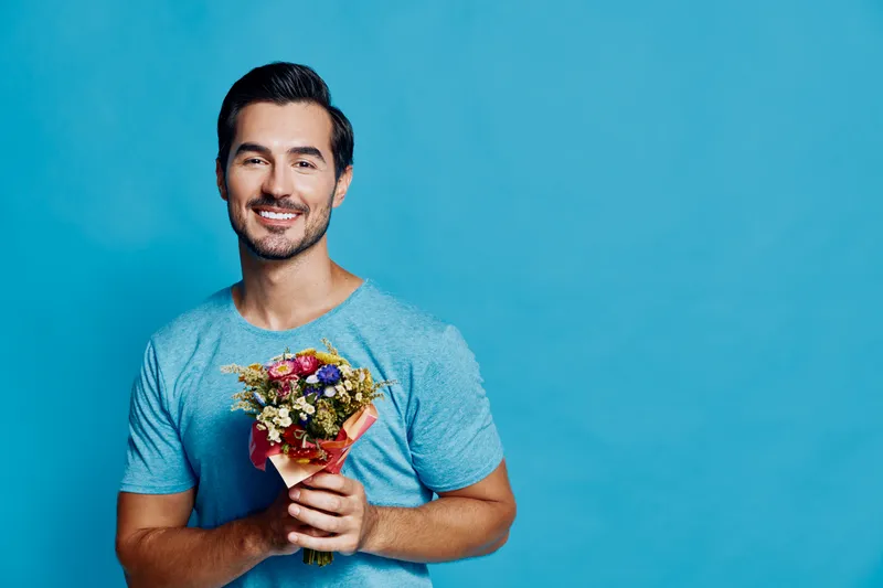 Happy smiling man holding a vibrant flower bouquet, wearing a blue t-shirt, standing against a bright turquoise background. Cheerful lifestyle concept.
