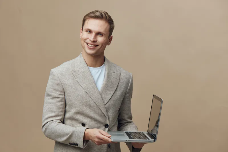 Smiling young man in smart casual blazer holding laptop computer, looking at camera, standing against plain beige background. Studio portrait, people, technology, casual style, positive mood, modern w
