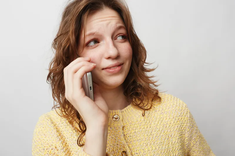 Young woman with wavy brown hair wearing yellow sweater talks on smartphone looking away with thoughtful expression. Isolated on white background. Concept of communication and casual lifestyle.