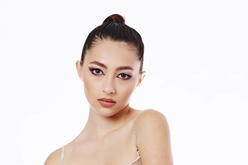 Young woman with artistic bright eye makeup and flawless skin poses confidently in studio on white background, expressing modern beauty and fashion creativity.