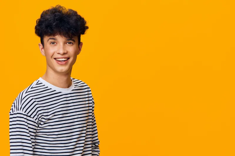 smiling young man with curly hair wearing striped shirt stands against vivid orange background, expressing joy and youthful energy in casual style.
