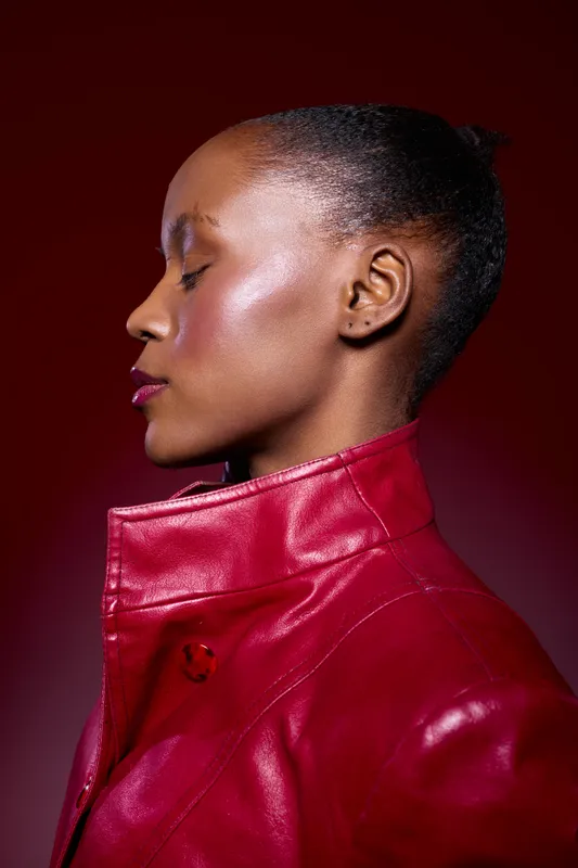 Profile portrait of a black woman with dark skin wearing a red leather jacket, eyes closed, against a dark red background. Concept of fashion, beauty, and confidence.