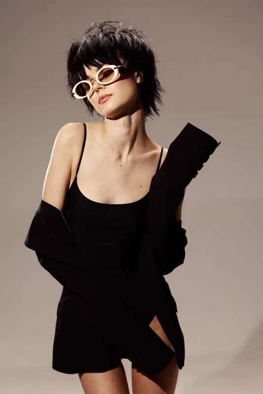 Stylish woman in black dress and sunglasses posing confidently with short hair and beige background.