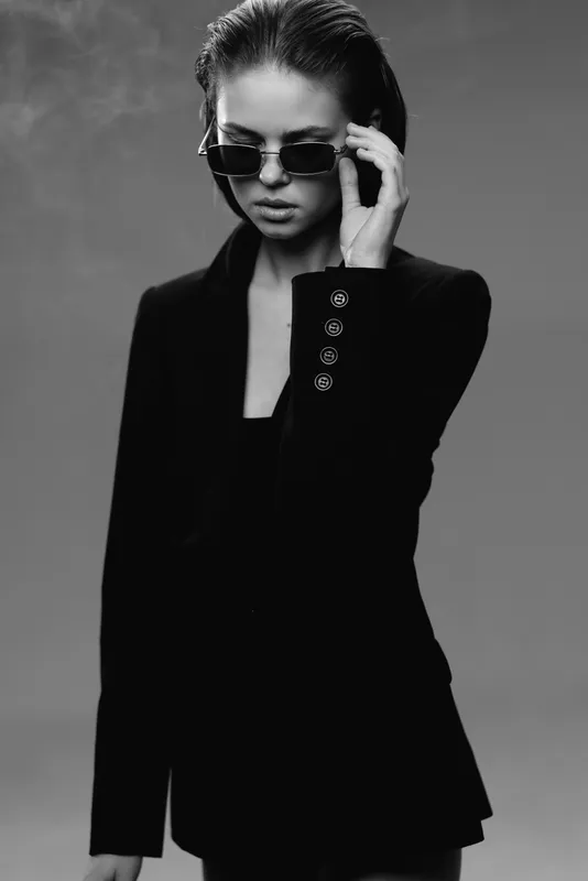 Black and white portrait of stylish woman wearing sunglasses and dark blazer with serious expression.
