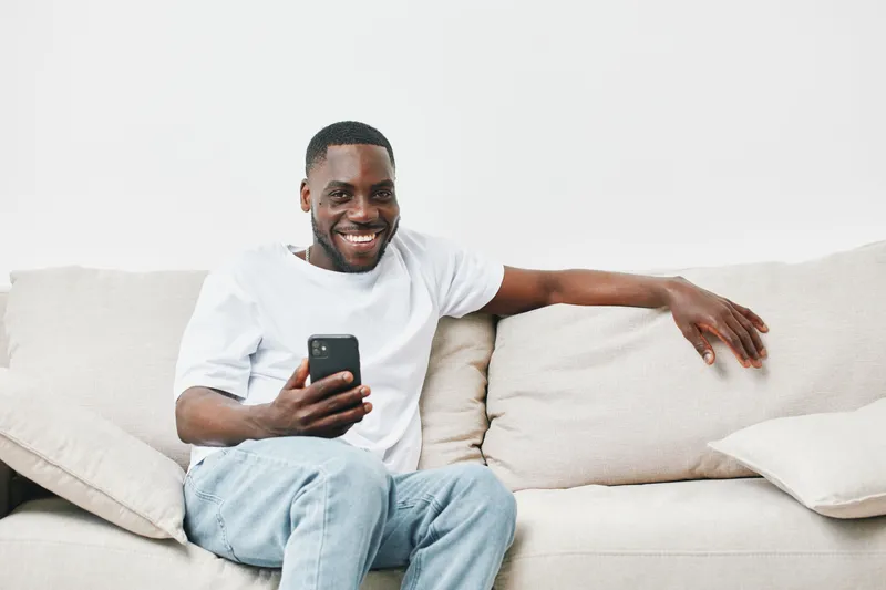 Cheerful African American man sitting on a couch using a smartphone, wearing casual clothing, smiling, light background, modern home interior, lifestyle concept