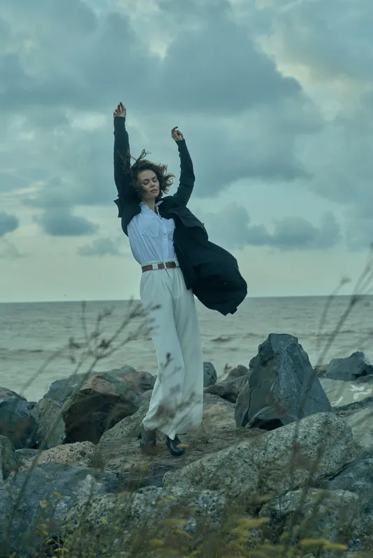 Woman leaps on a rocky shoreline, wind lifting a long coat as waves crash nearby, creating a dynamic outdoor scene of strength, freedom, and motion by the sea.