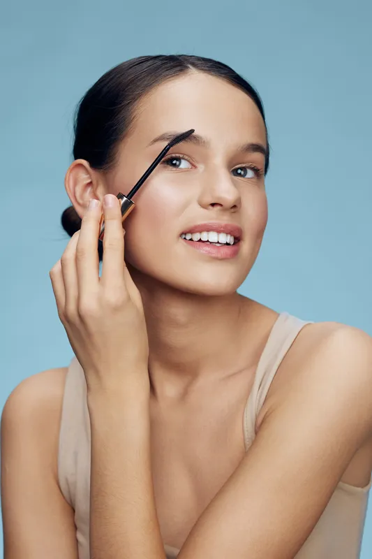 A young woman applying eyebrow gel, showcasing a natural beauty look against a soft blue background, highlighting beauty, makeup, and self care concepts