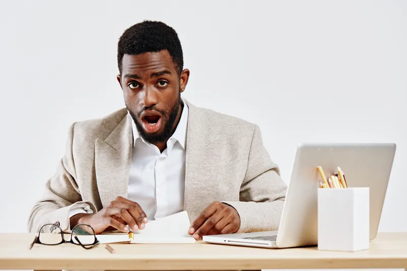 Excited young black man in beige blazer and white shirt sitting at a desk with open laptop and stationery, expressing surprise or shock. Modern office environment, casual professional style. People em