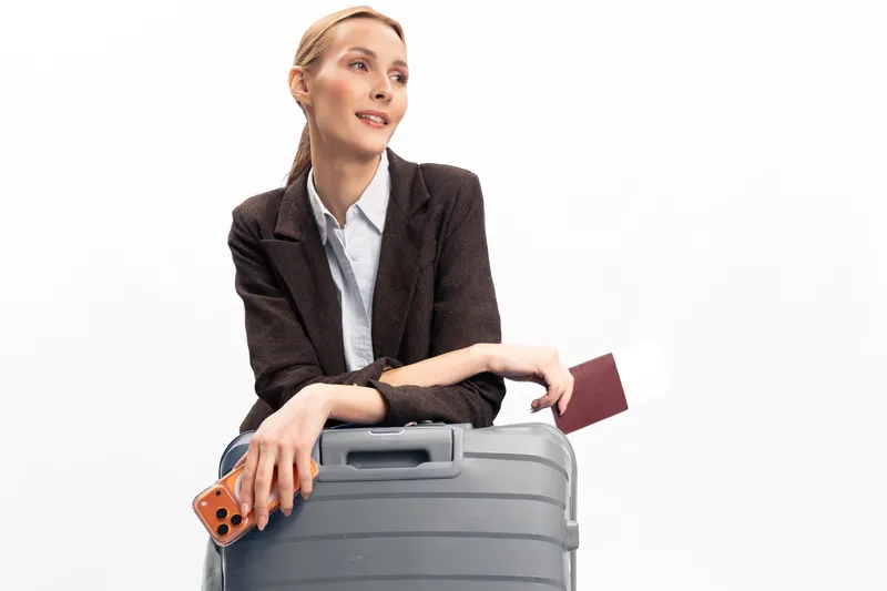 Young woman with suitcase holds passport and smartphone, looking away with a smile. Concept for travel, vacation, tourism, and holiday preparation on white background.